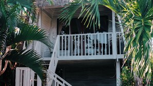 Een huis met twee verdiepingen, voorzien van een rieten dak, een wit balkon en een trap die naar de bovenverdieping leidt.
