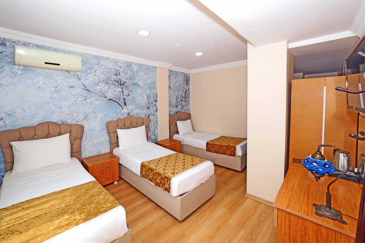 A hotel room with three beds, a wooden floor, a wall-mounted air conditioner, and a small table with a lamp.