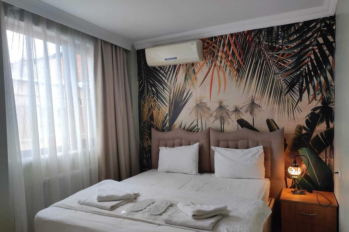 A hotel room with a bed, a nightstand, an air conditioning unit, and a tropical-themed wall mural.