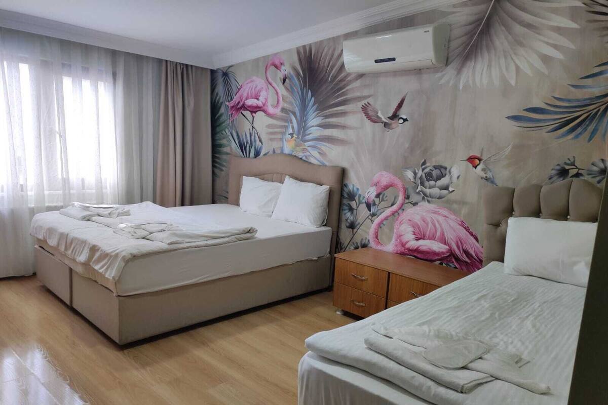 A hotel room with two beds, a wooden floor, a large window with sheer curtains, and a wall mural featuring flamingos and birds.