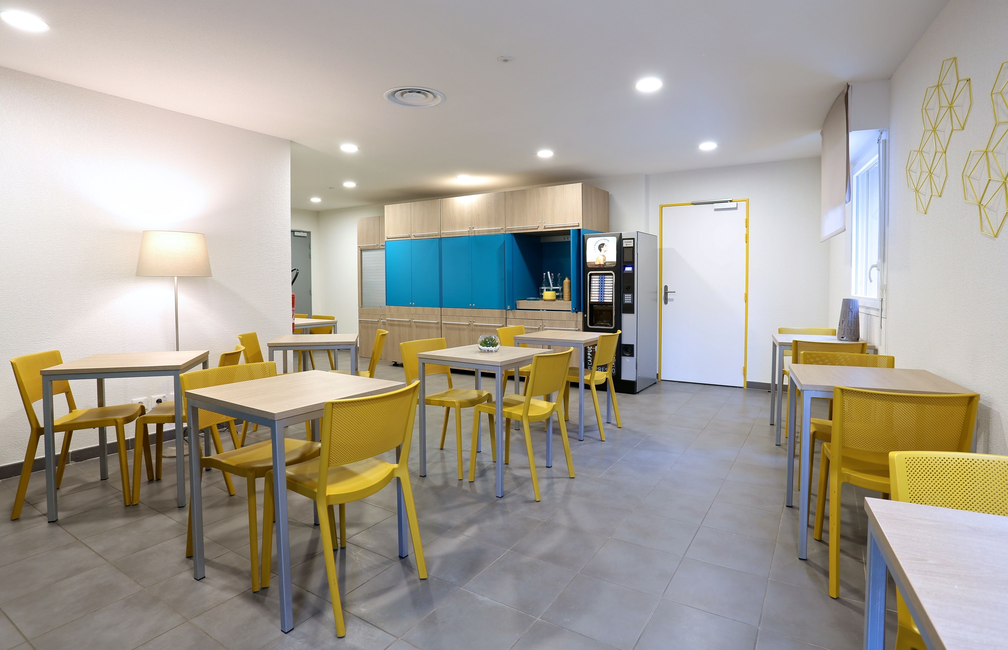 Een moderne eethoek met gele stoelen en tafels, een keuken op de achtergrond en een licht plafond.