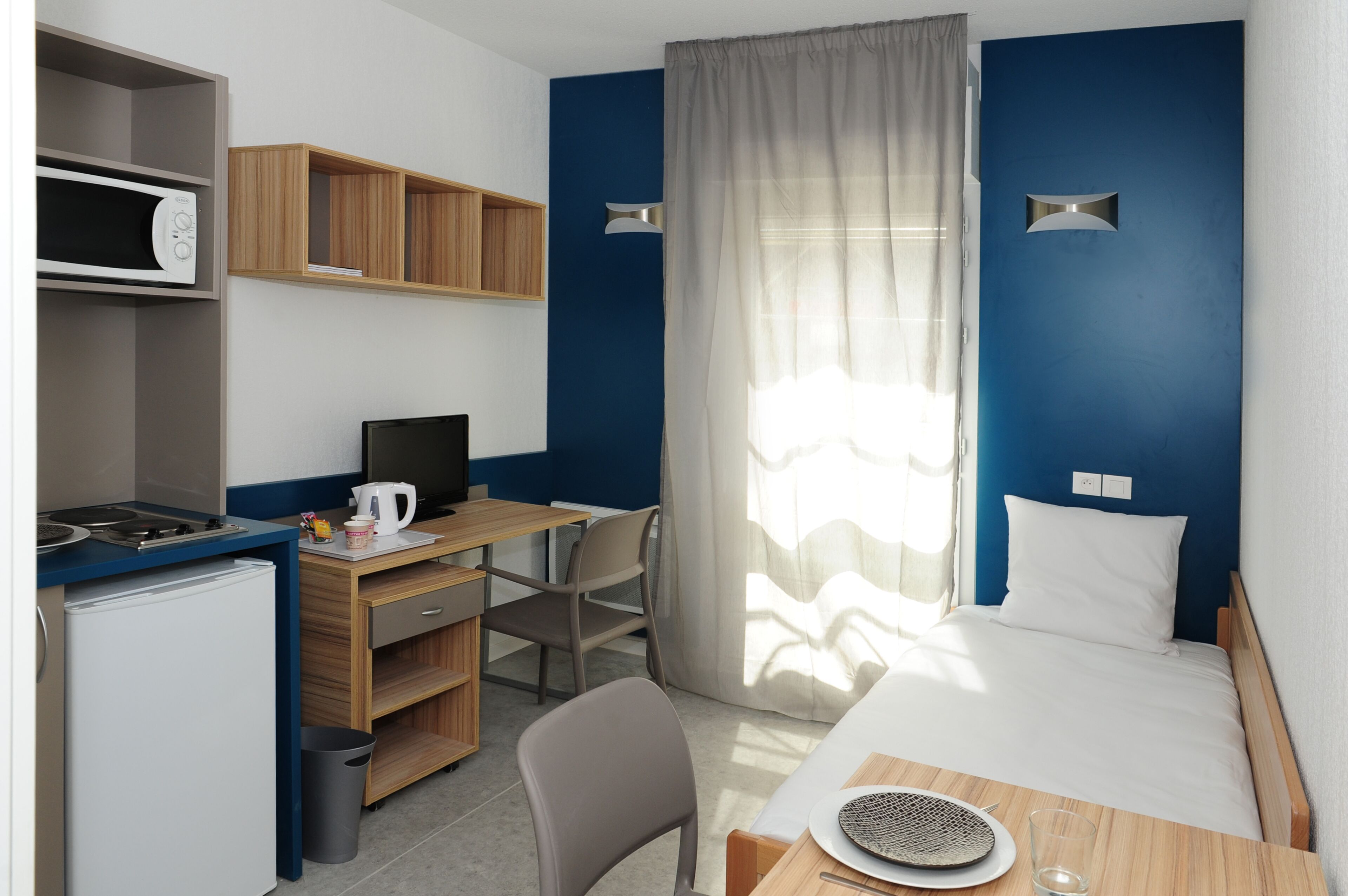 Une chambre d’hôtel compacte comprenant un coin cuisine, un bureau, un lit et une grande fenêtre avec des rideaux légers.