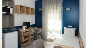 Une chambre d’hôtel compacte comprenant un coin cuisine, un bureau, un lit et une grande fenêtre avec des rideaux légers.