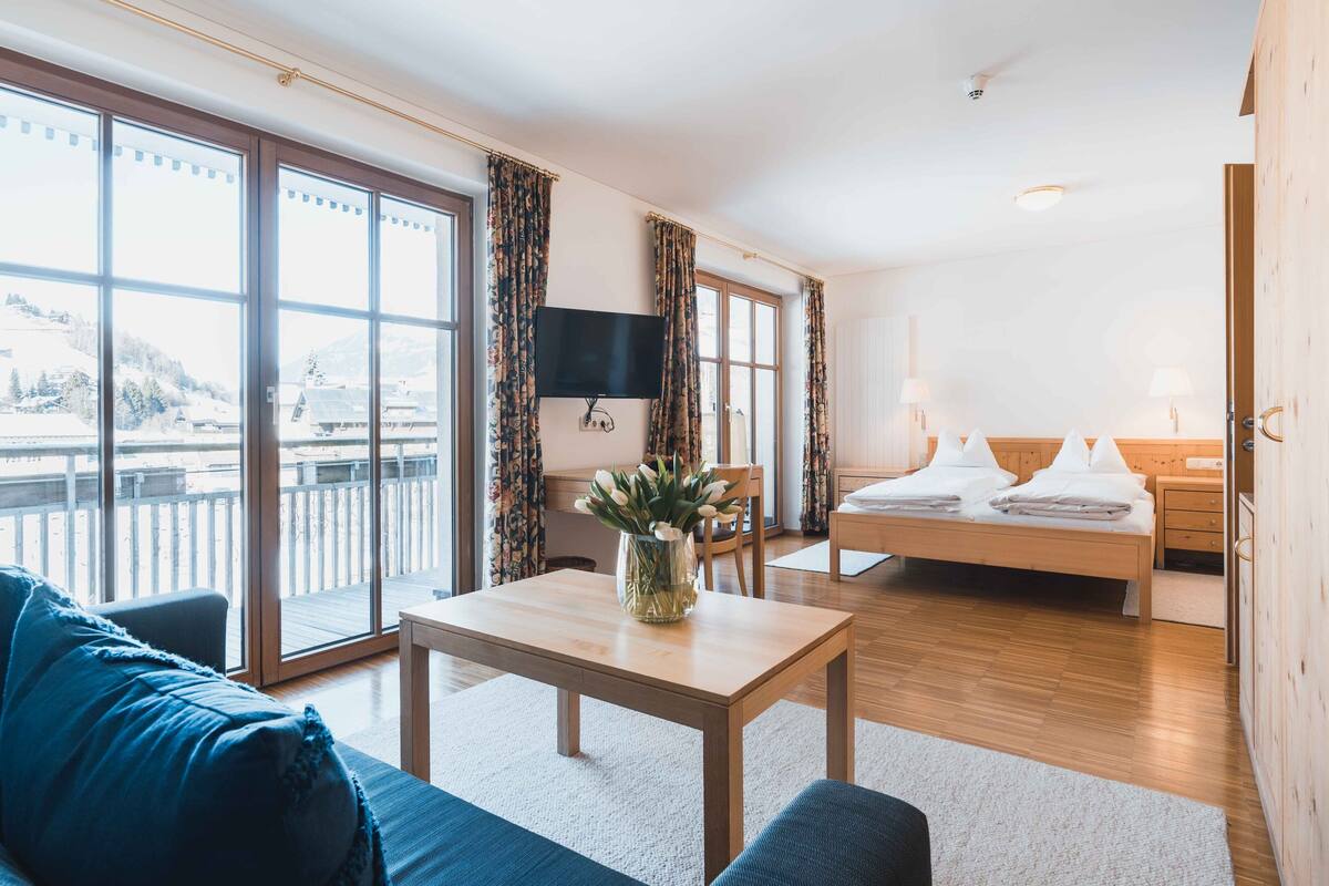 A modern hotel room with a large sliding glass door to a balcony, a bed with white linens, a flat-screen TV, and a small table with a vase of flowers.