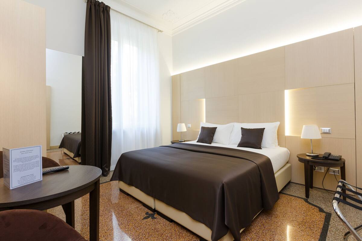 A hotel room with a bed, bedside tables, a desk, a chair, and a window with curtains.