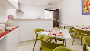 A modern dining area with green chairs and white tables, featuring a kitchen counter with food items and a colorful painting on the wall. - Domus Terenzio (Rome)
