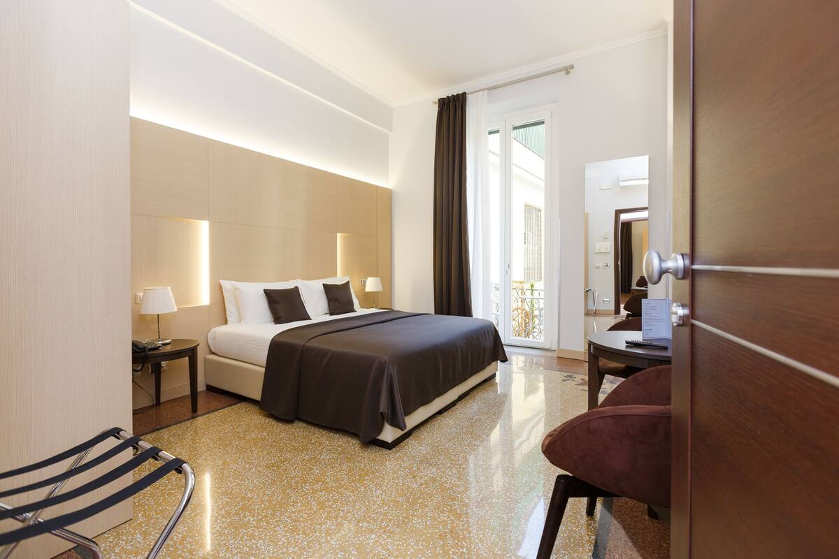 A hotel room with a bed, a desk, a chair, a lamp, and a view of the outside.