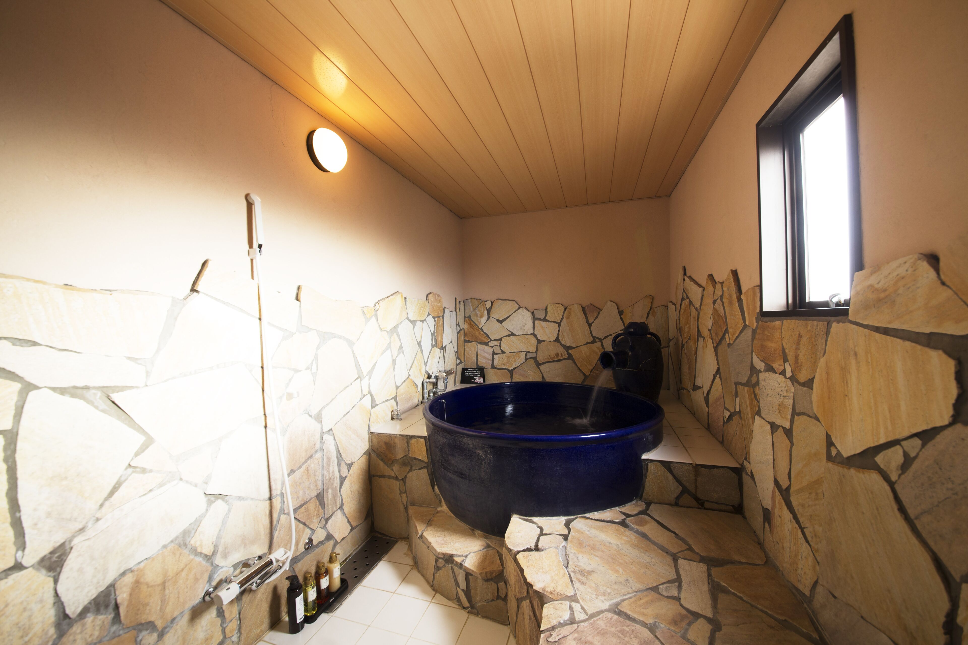 A stone-walled bathroom with a large blue bathtub, a wooden ceiling, and a window.