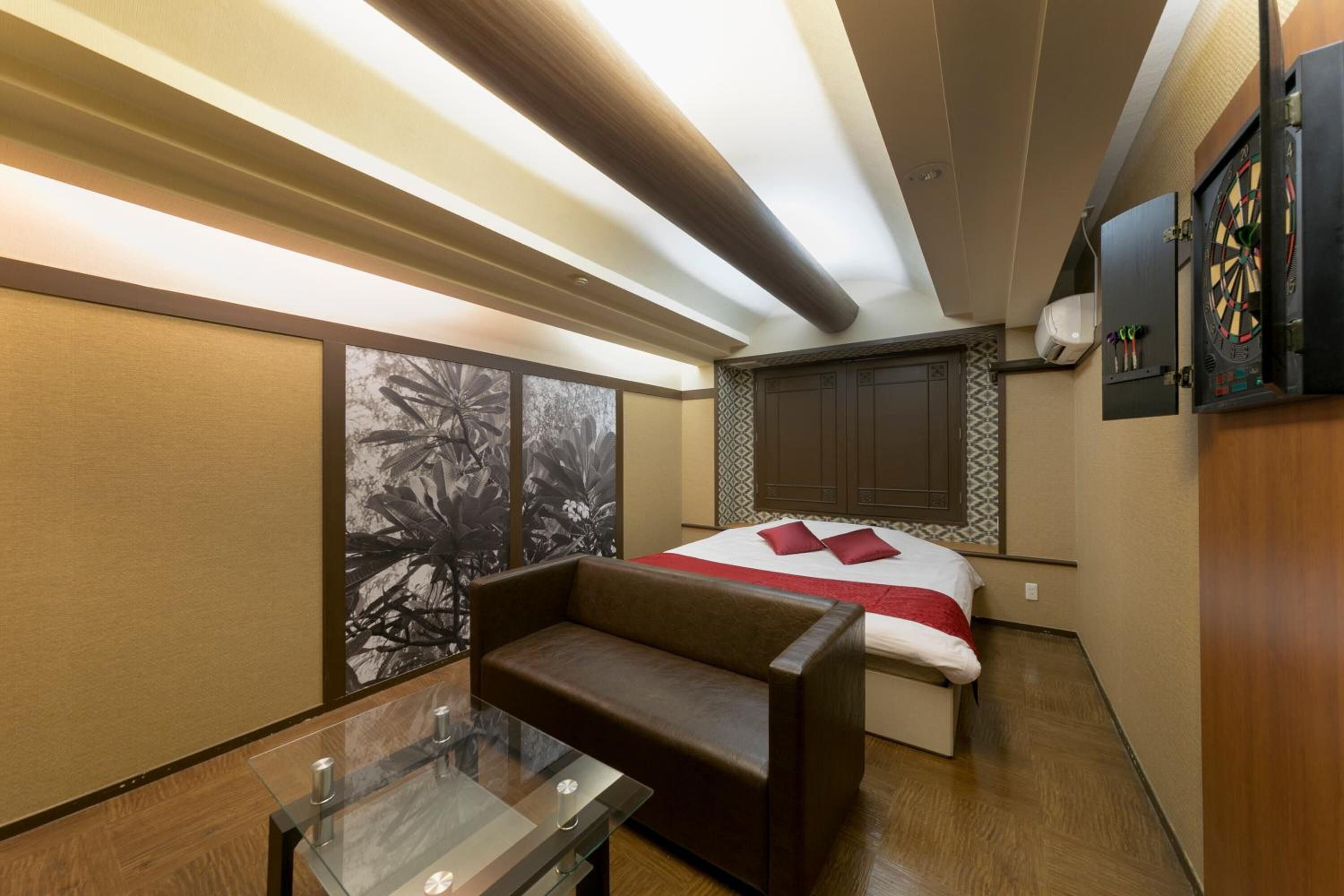 A compact hotel room with a bed, a sofa, a glass table, and a dartboard on the wall.