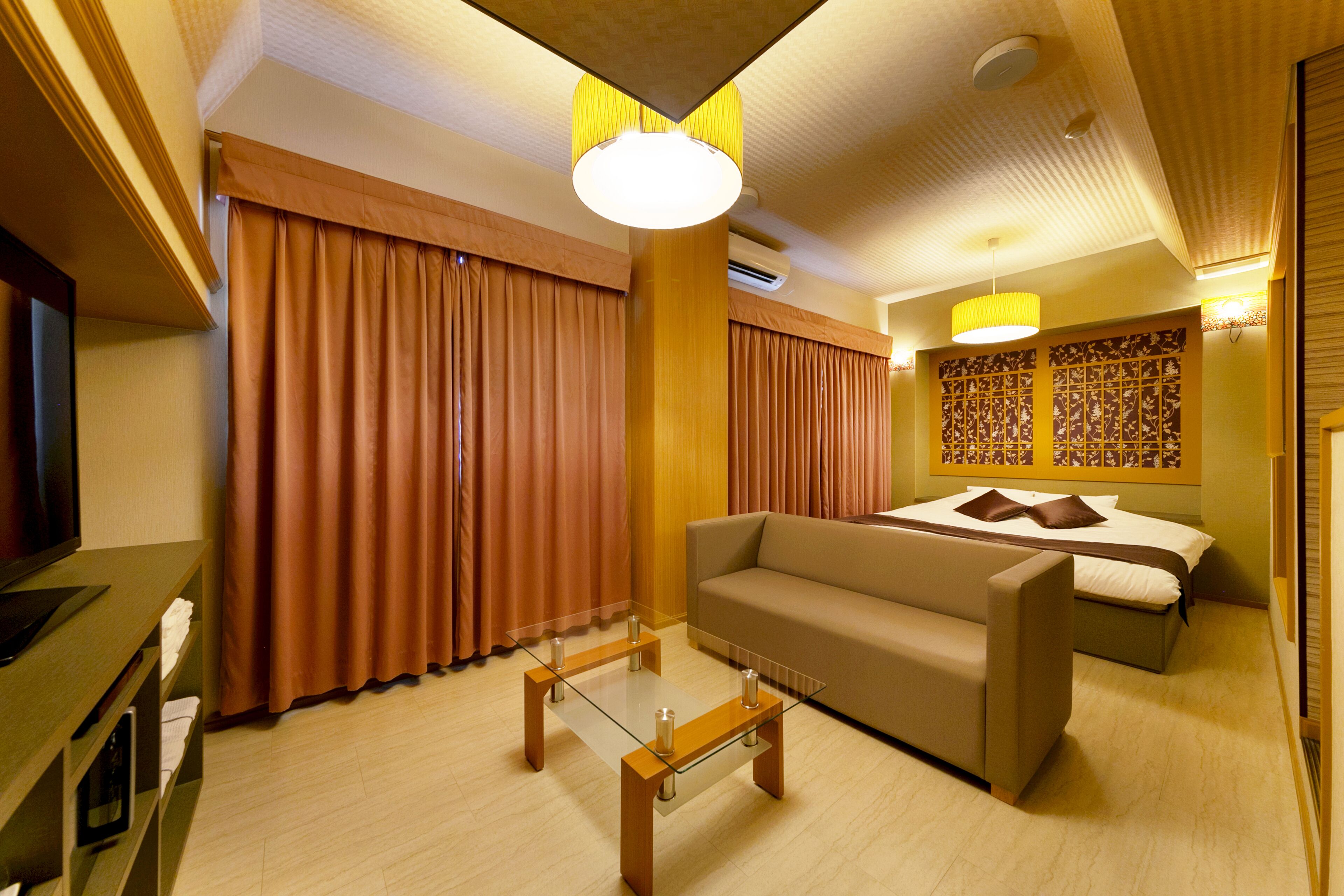 A hotel room with a bed, sofa, glass coffee table, curtains, and a television.