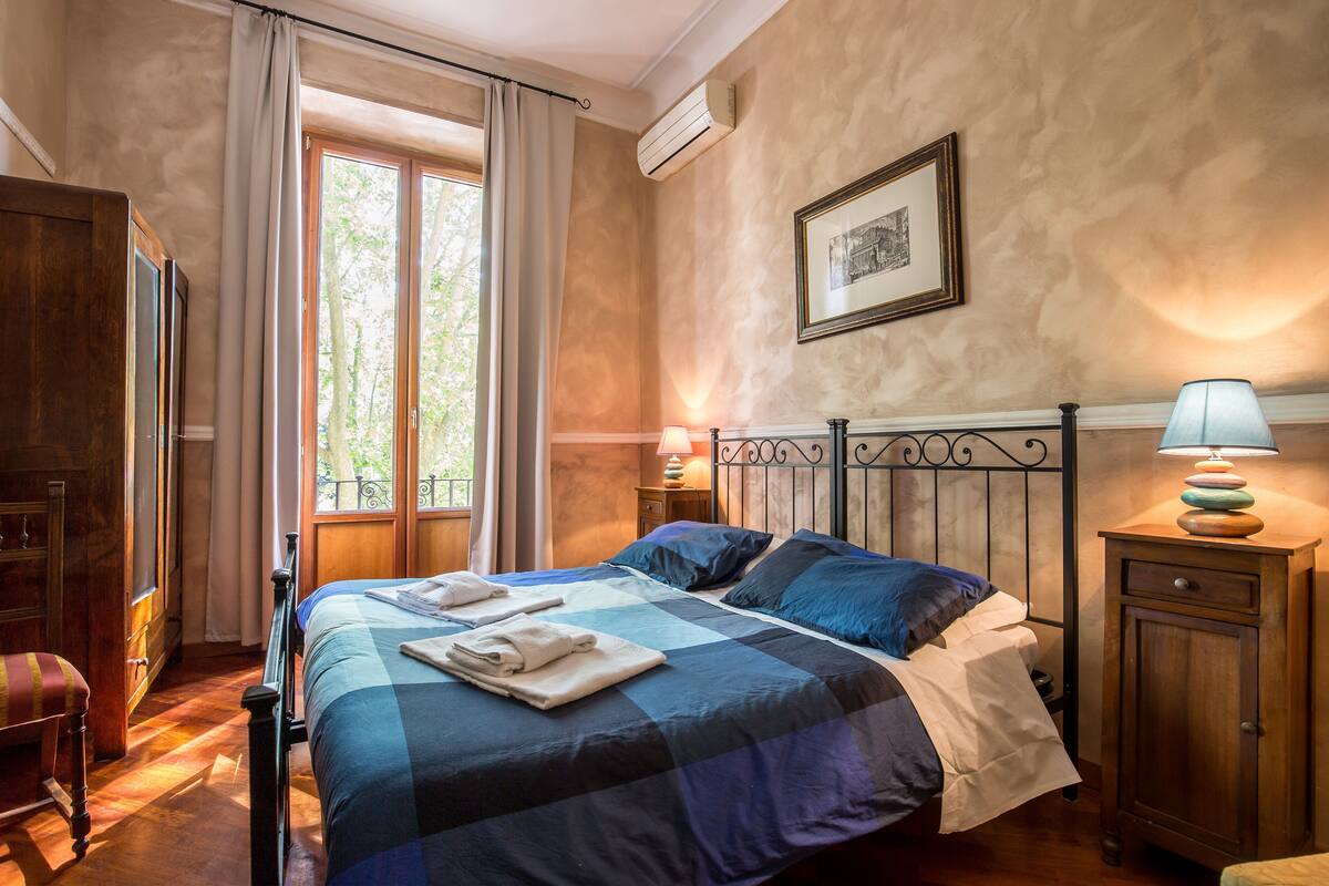 A bedroom with a checkered bedspread, a wooden nightstand with a lamp, and a window with curtains.