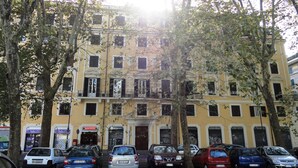 Un edificio amarillo de varios pisos con balcones, autos estacionados al frente y árboles en primer plano.