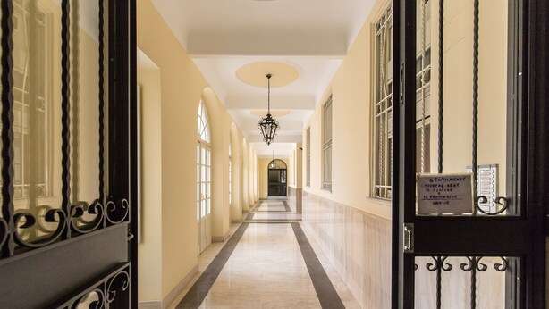Un largo pasillo con piso de mármol, ventanas grandes y un candelabro.