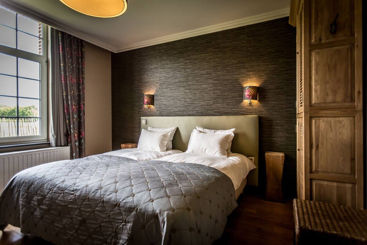 A bedroom with a large bed, two bedside tables, a wooden headboard, and a window with curtains.