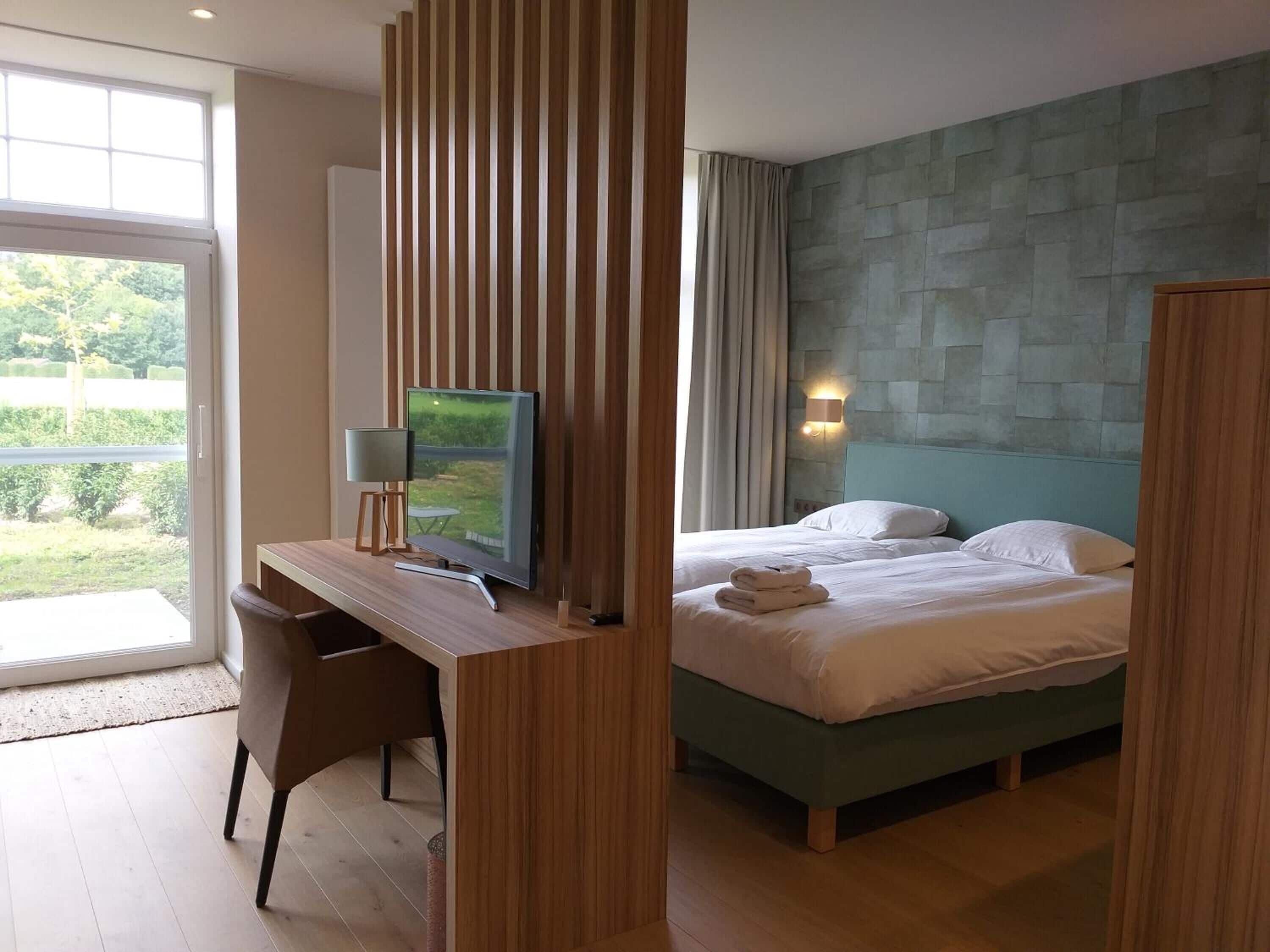 Una habitación de hotel moderna con escritorio, televisión de pantalla plana, cama con dos almohadas y vistas a zonas verdes por la ventana.