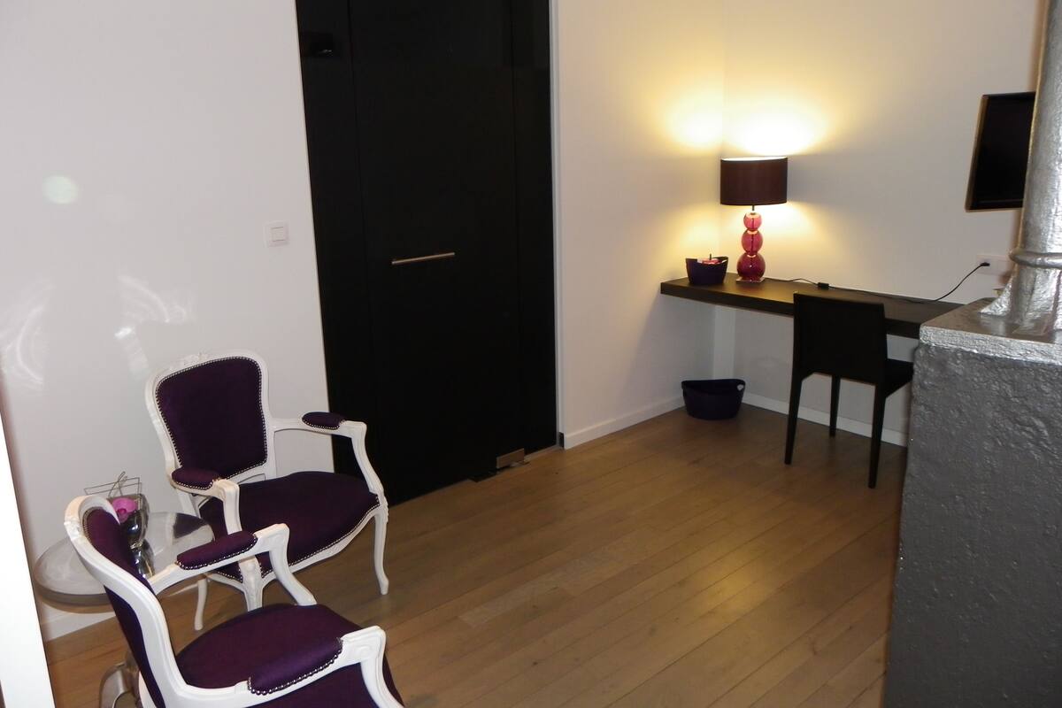 A small, compact room with a wooden floor, a black door, a desk with a lamp, and two purple chairs.
