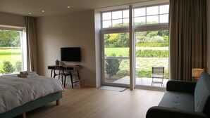 A bedroom with a bed, a desk with a TV, a sofa, and a large sliding glass door leading to a garden. - Boskapelhoeve Charming Hotel (Buggenhout)