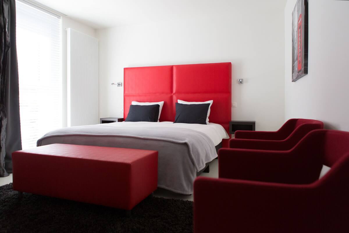 A modern bedroom with a red headboard, two black and white pillows, a grey bedspread, a red bench, and a black rug.