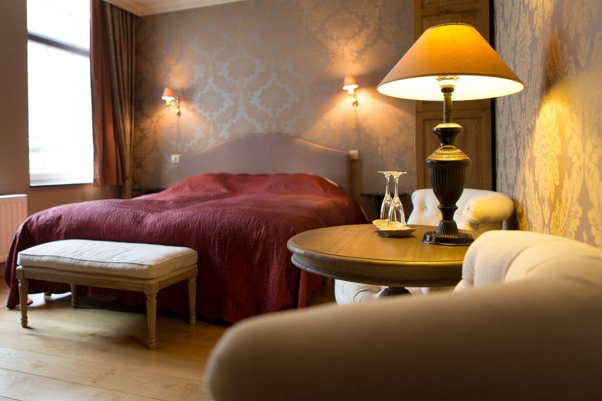 A hotel room with a bed, a bedside table with a lamp, a chair, and a window with curtains.