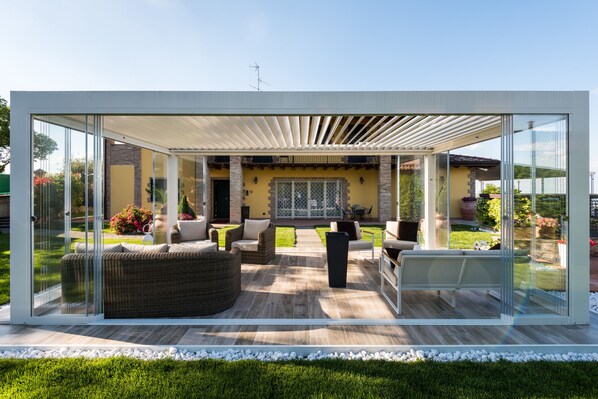 A modern outdoor patio with glass walls, wicker furniture, and a view of the garden.
