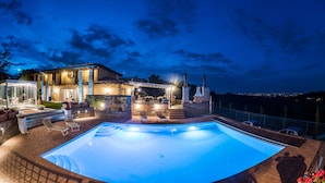 Ein Swimmingpool mit Liegestühlen, einer überdachten Terrasse und Blick auf die nächtliche Stadt.