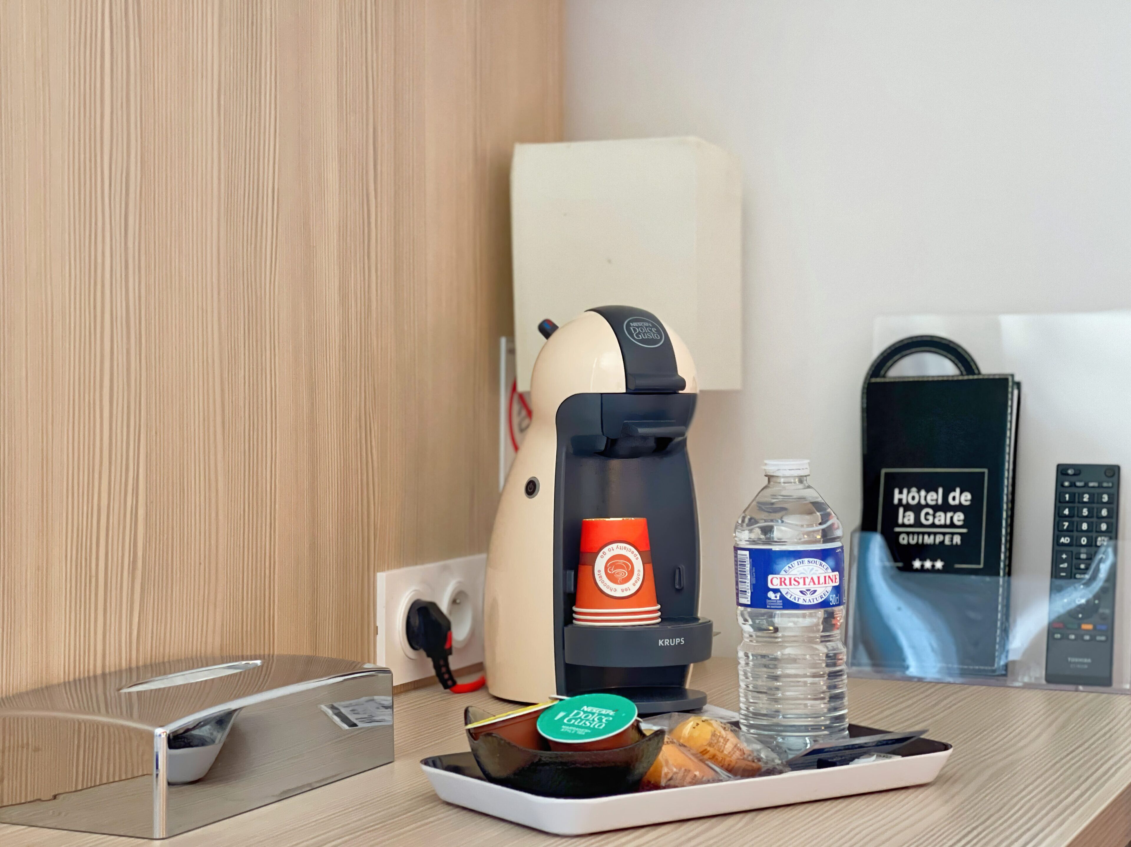 A hotel room with a coffee machine, a water bottle, a remote control, and a tray with food items.