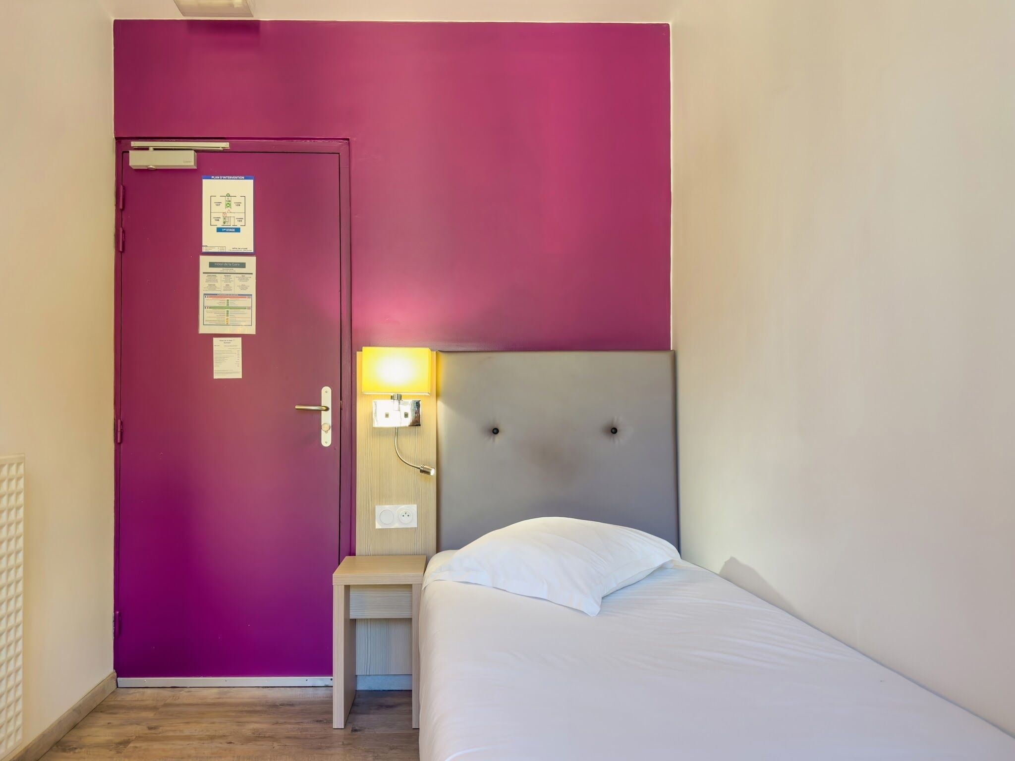 A room with a purple door, a bed with a white headboard, and a small bedside table with a lamp.