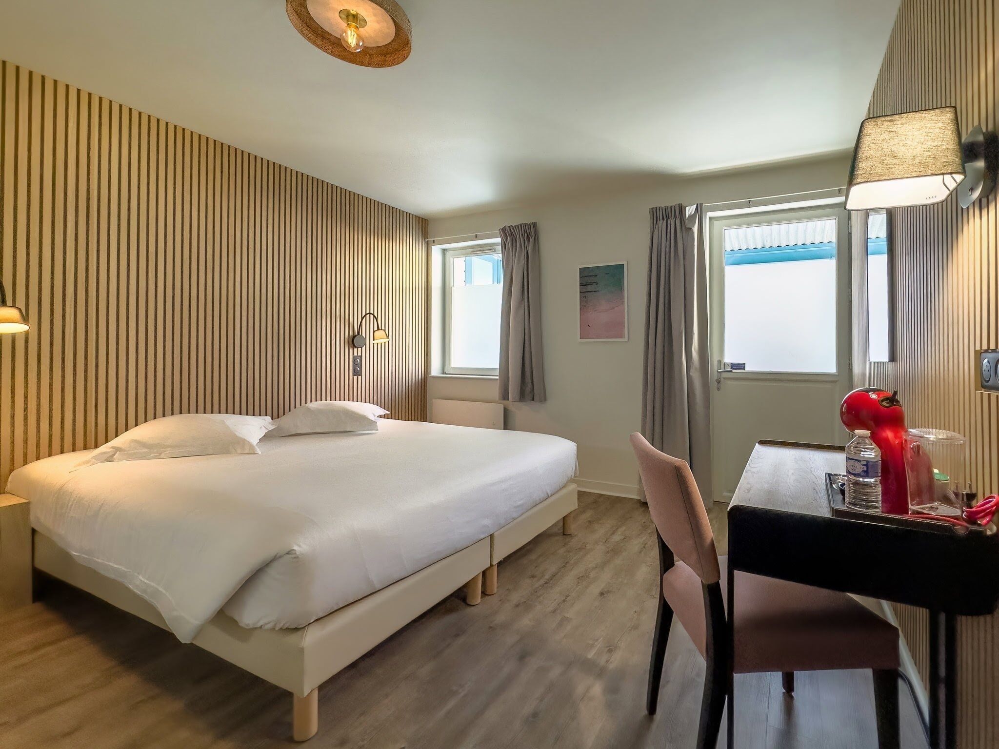 Une chambre d’hôtel avec un lit, un bureau avec une chaise, une lampe et une fenêtre avec des rideaux.