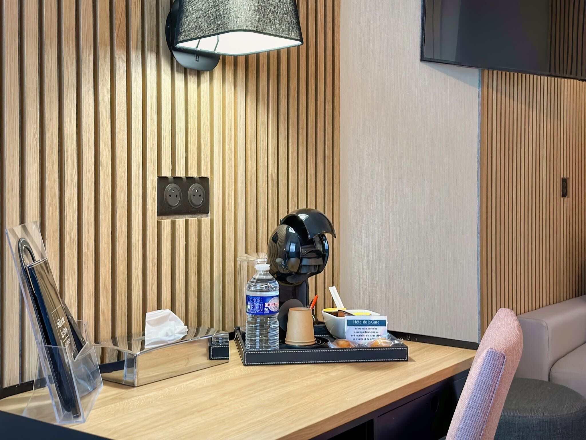 A hotel reception desk with a wooden panel wall, a mounted lamp, a telephone, a water bottle, a coffee cup, a small tray, and a sign indicating the hotel name.