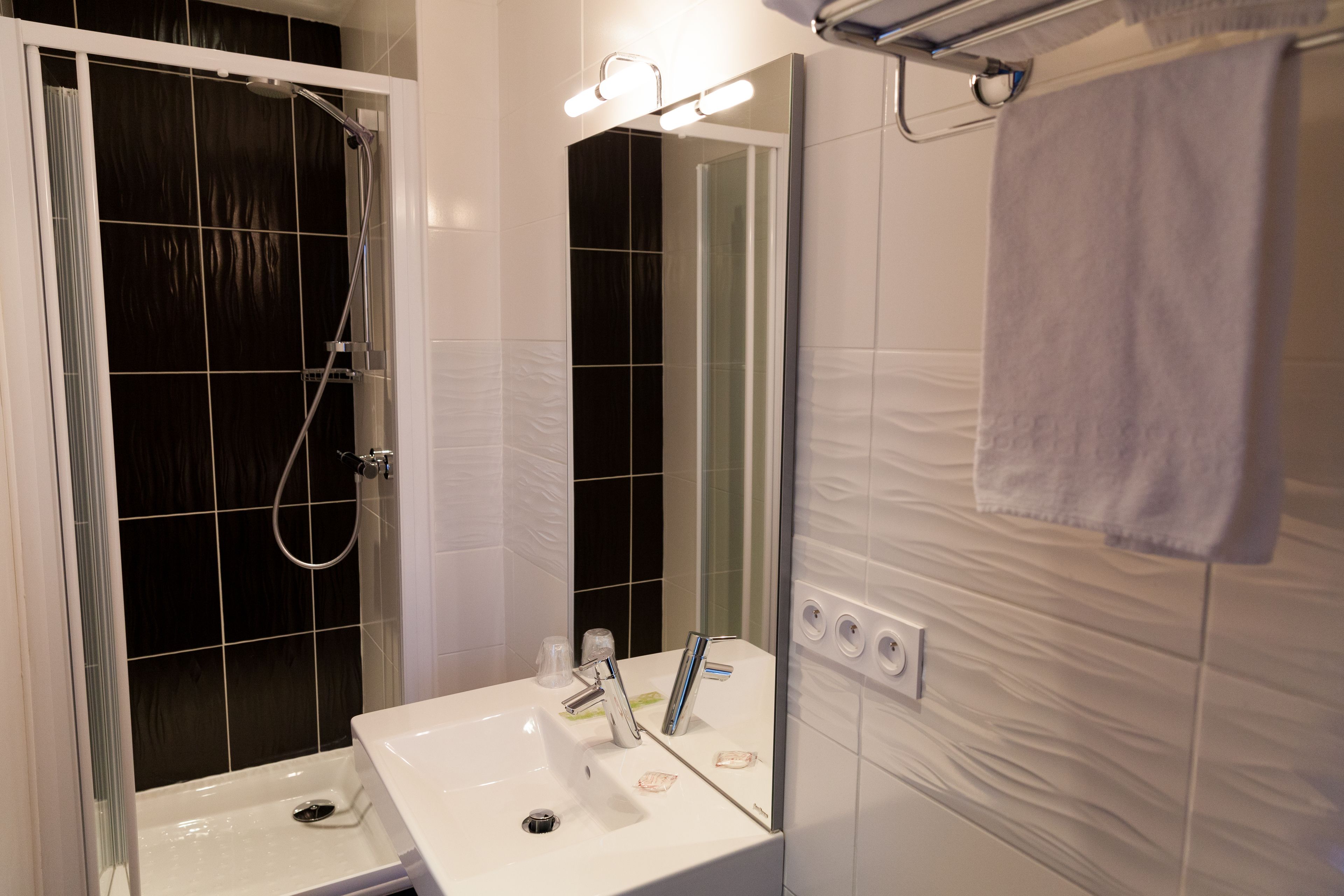 Un baño con un lavamanos blanco, un espejo, una ducha con puerta de vidrio y una toalla colgada en un perchero.