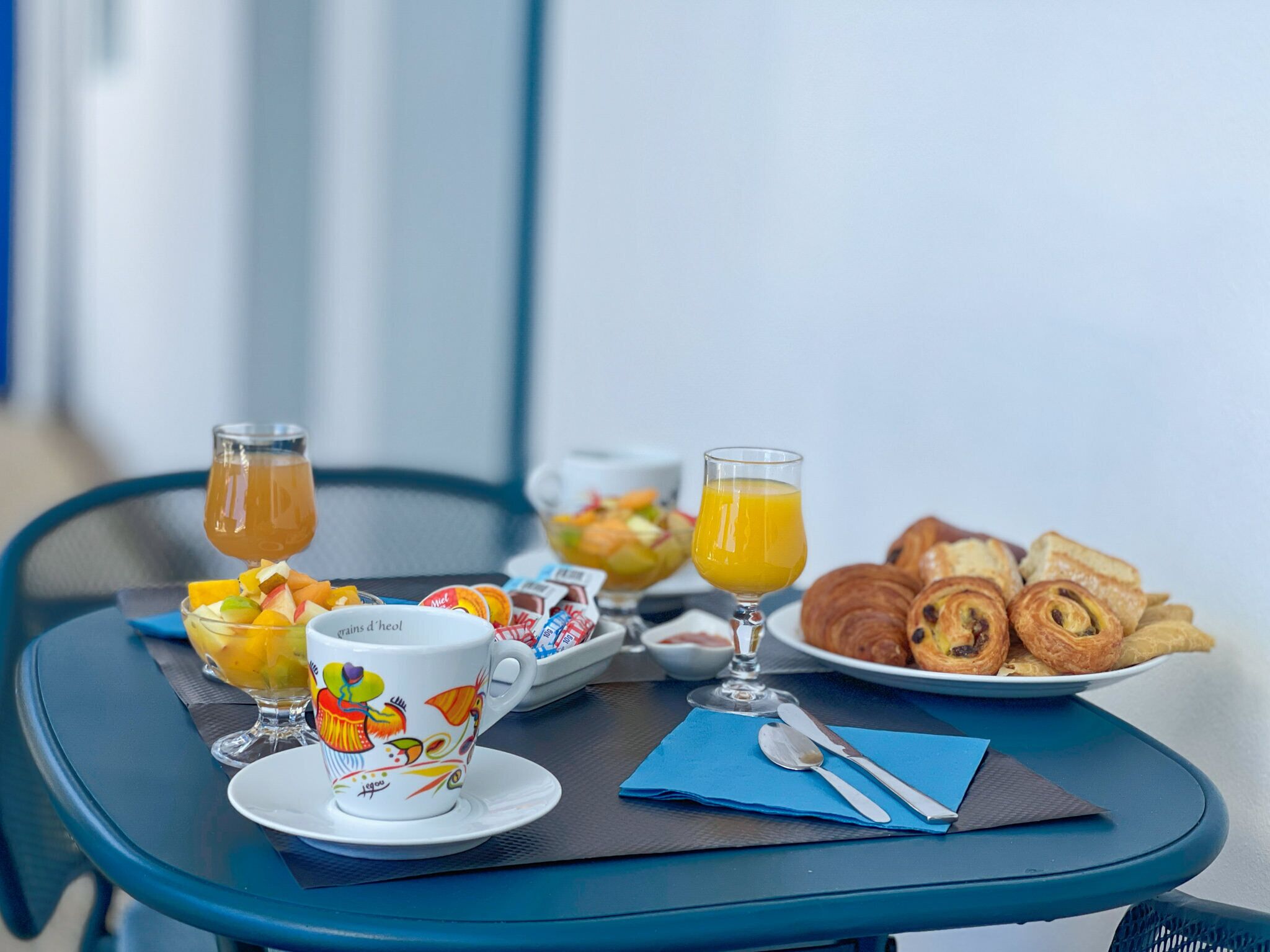 A breakfast table with a cup, two glasses of orange juice, a bowl of fruit, croissants, and a plate of pastries.