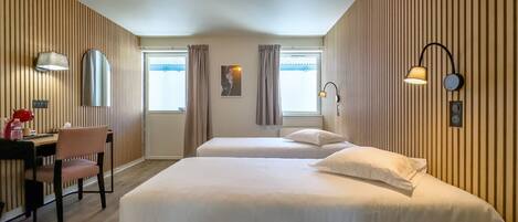 A hotel room with two beds, a desk with a chair, a lamp, and a wall-mounted light fixture.