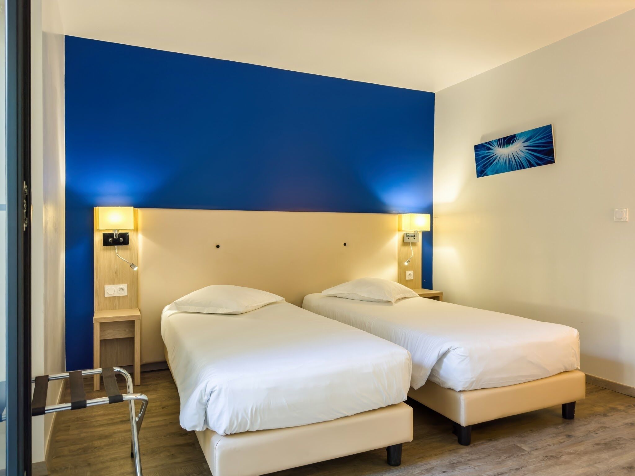 Een hotelkamer met twee bedden, een houten nachtkastje, een wandlamp en een blauw abstract schilderij.