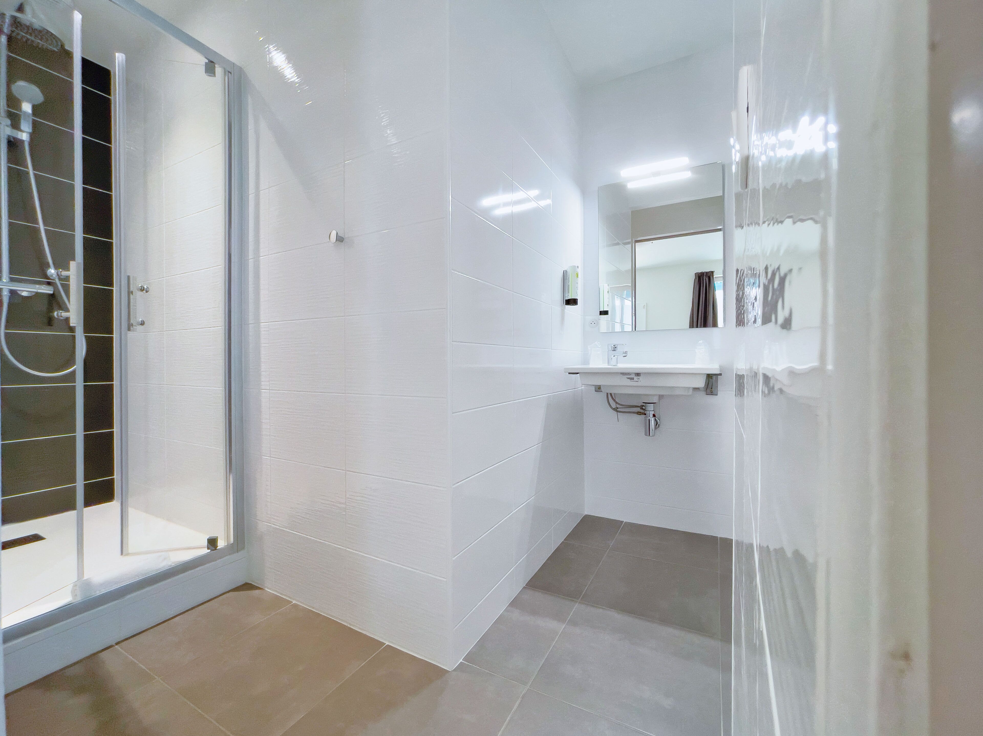 Une salle de bain avec des murs recouverts de carreaux blancs, une douche en verre et un lavabo avec un miroir au-dessus.