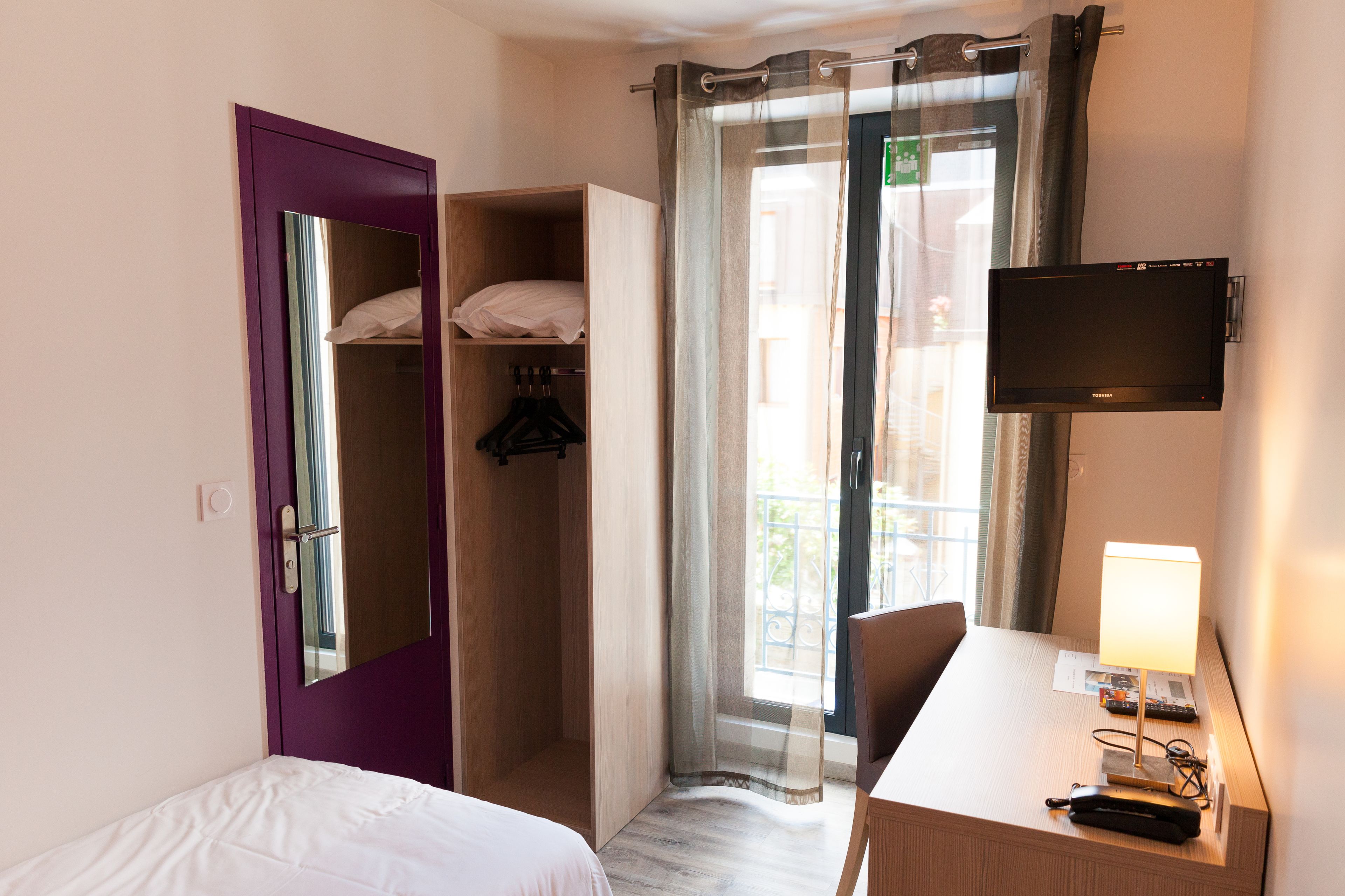 A hotel room with a bed, a desk with a lamp, a TV, a wardrobe, and a window with curtains.