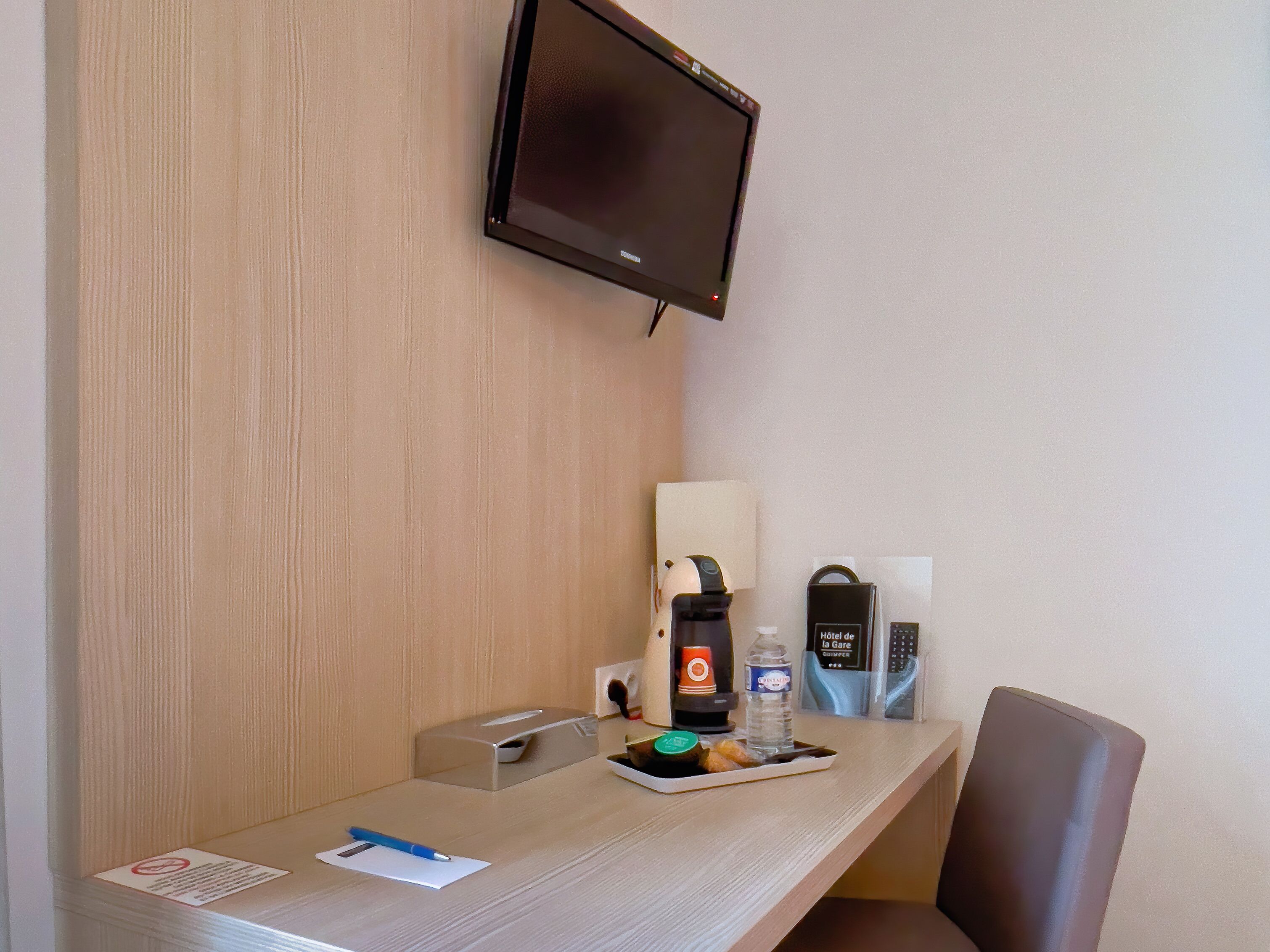 A hotel room with a flat-screen TV mounted on the wall, a desk with a notepad, pen, and a tray with a bottle and snacks.