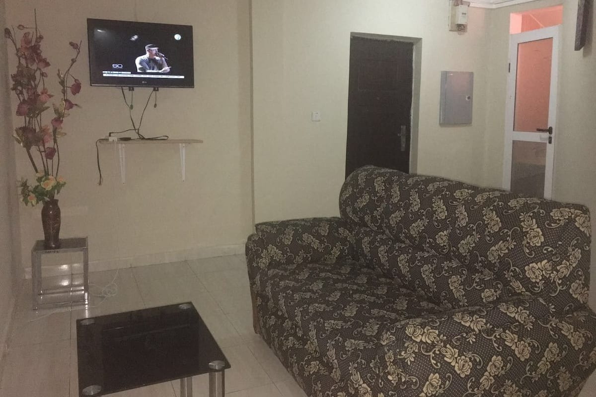 A living room with a sofa, a TV mounted on the wall, a small table, and a vase with flowers.