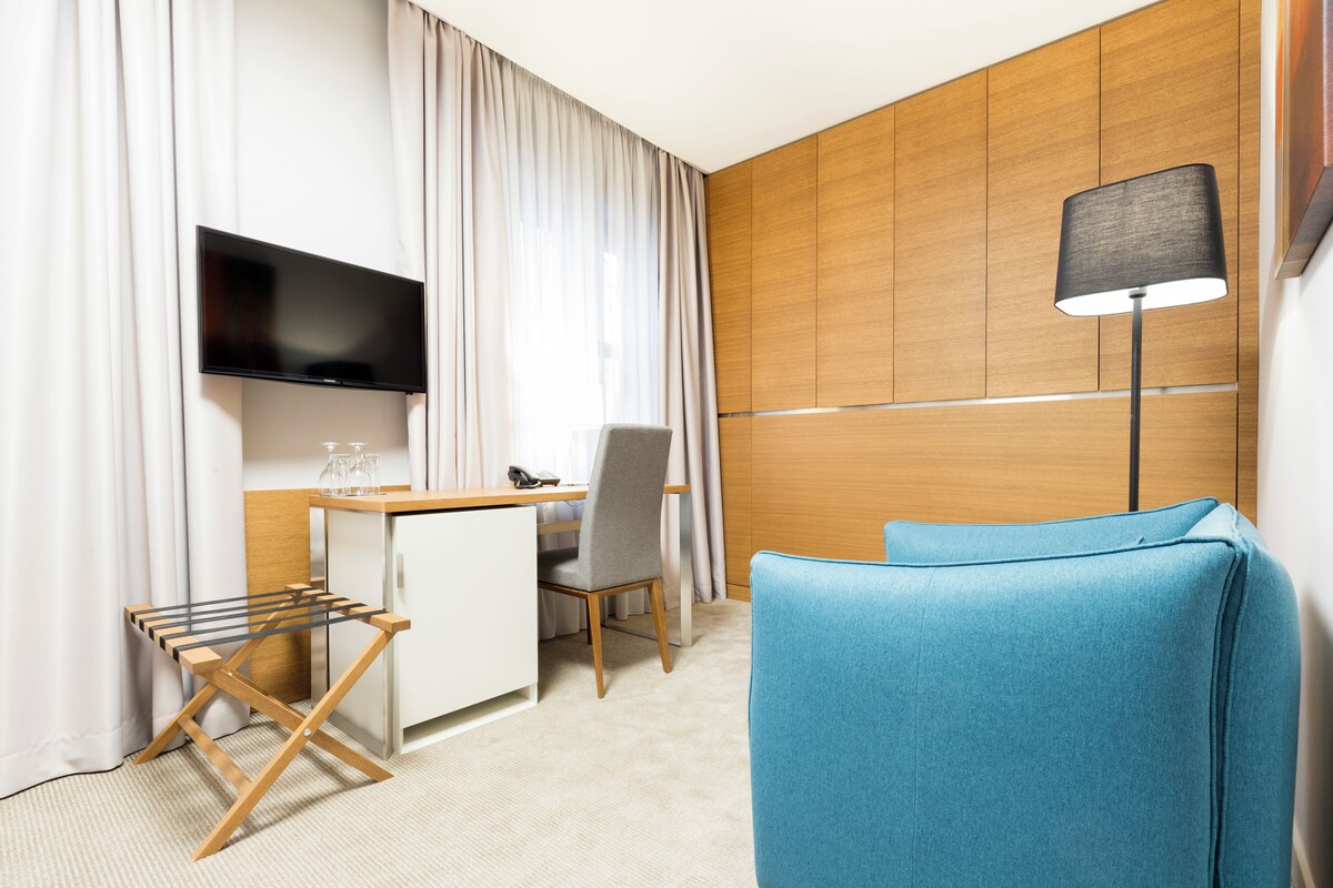 Habitación de hotel con televisor montado en la pared, un escritorio con silla, un sofá azul y una lámpara de pie.