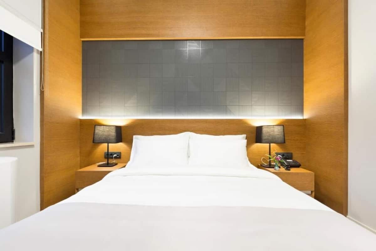 Una habitación de hotel con una cama, dos mesitas de noche con lámparas, un cabecero y un ventanal.