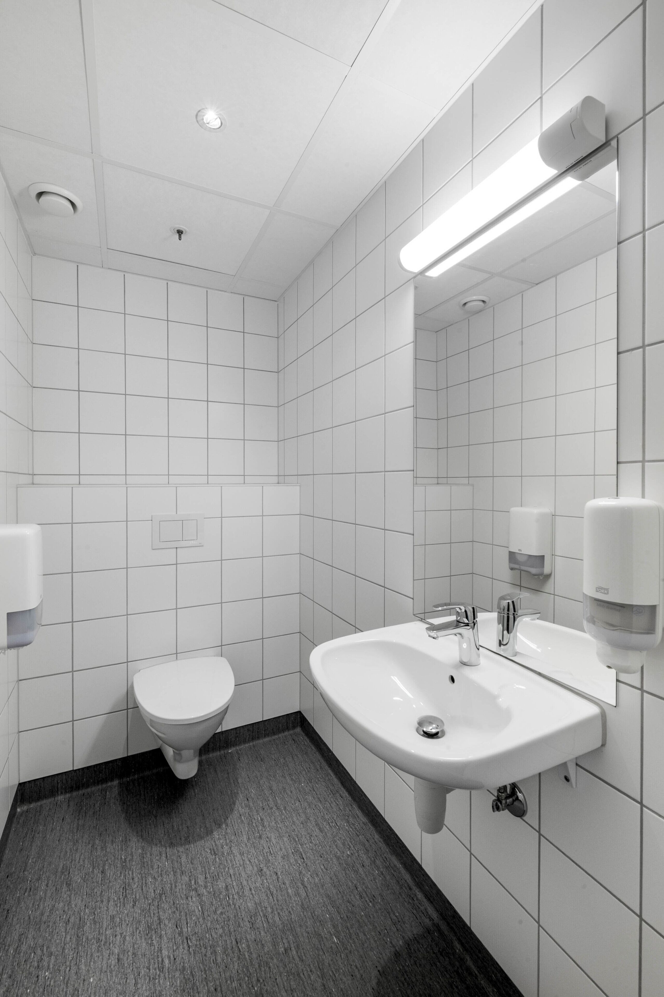 Un baño moderno con azulejos blancos, un inodoro, un lavamanos con espejo y un secamanos.