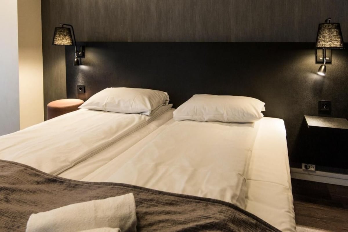 Una cama doble con sábanas blancas, dos lámparas fijadas a la pared, un cabecero de madera y un pequeño taburete de madera.