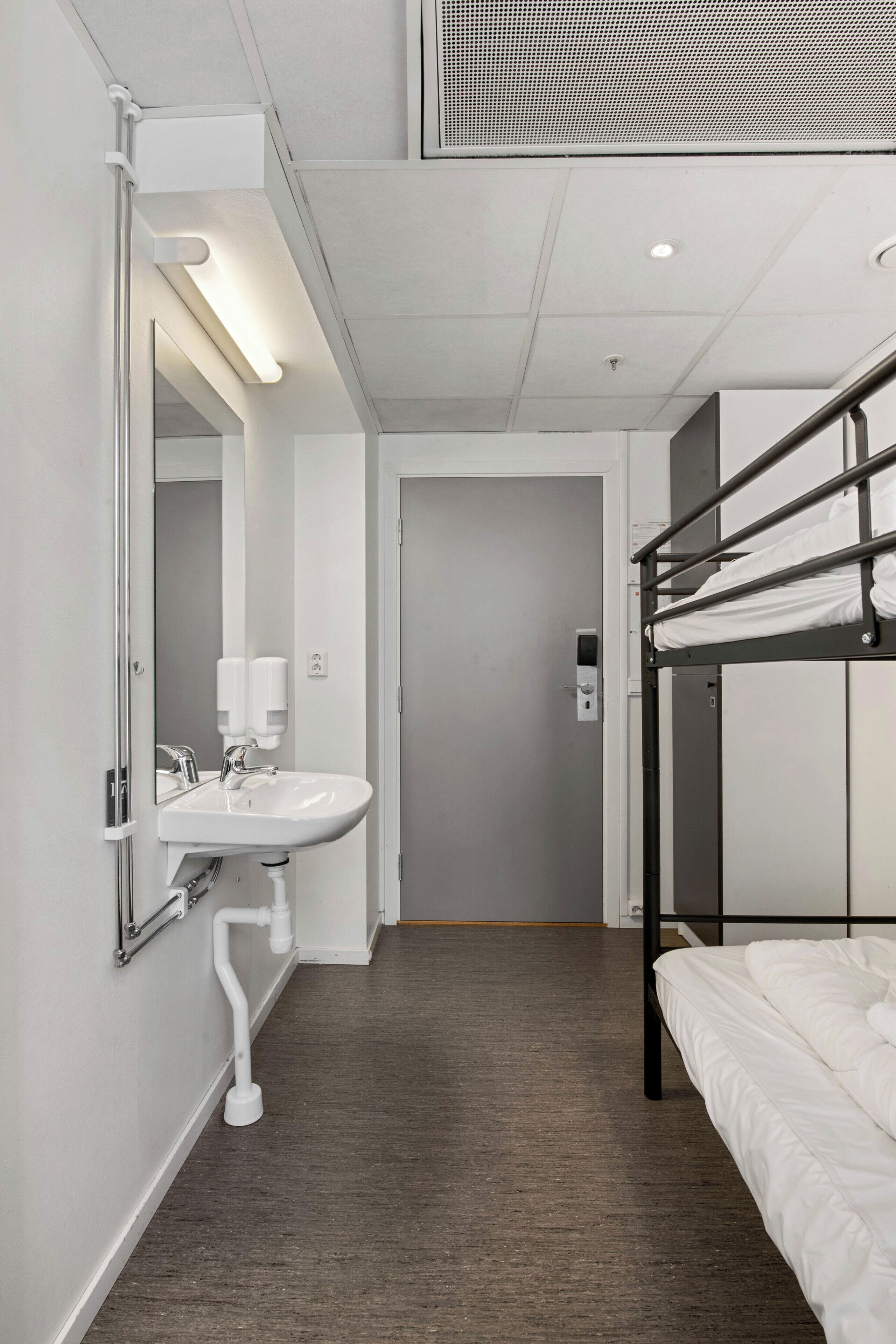 Une chambre d’hôtel avec un lit superposé, un lavabo blanc avec un miroir et une porte donnant sur une autre pièce.