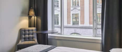 A hotel room with a bed, a chair, a table, and a window with a view of a building.