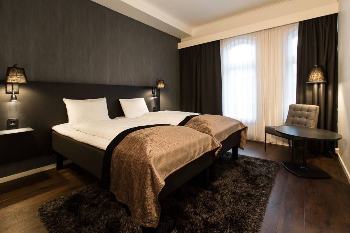 Quarto de hotel com duas camas, paredes escuras, uma janela grande com cortinas, uma mesa redonda pequena e duas luminárias fixadas na parede.