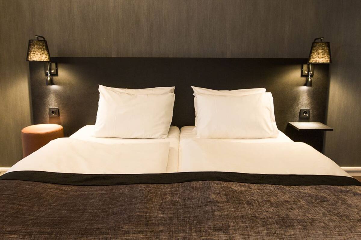 Cama bem arrumada com lençóis brancos e manta marrom escura. Há duas luminárias fixadas na parede, uma de cada lado da cama.