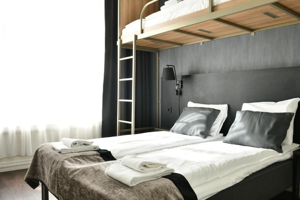 A modern hotel room with a bunk bed, a bedside table with a lamp, and a window with curtains.