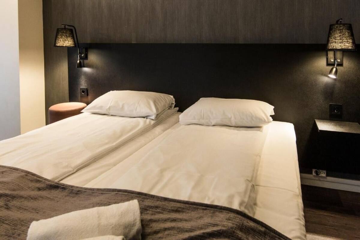 Uma cama de casal com lençóis brancos, duas luminárias fixadas na parede, uma cabeceira de madeira e um banquinho pequeno de madeira.