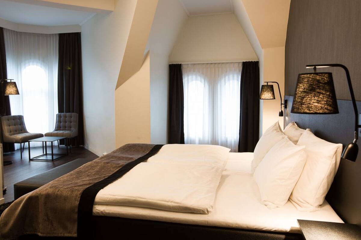 Quarto de hotel com uma cama grande, duas luminárias de cabeceira, uma cadeira e vista para o exterior pela janela.
