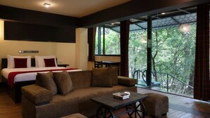 Habitación de hotel moderna con una cama grande, un sofá, una mesita y un balcón con vistas a árboles.
