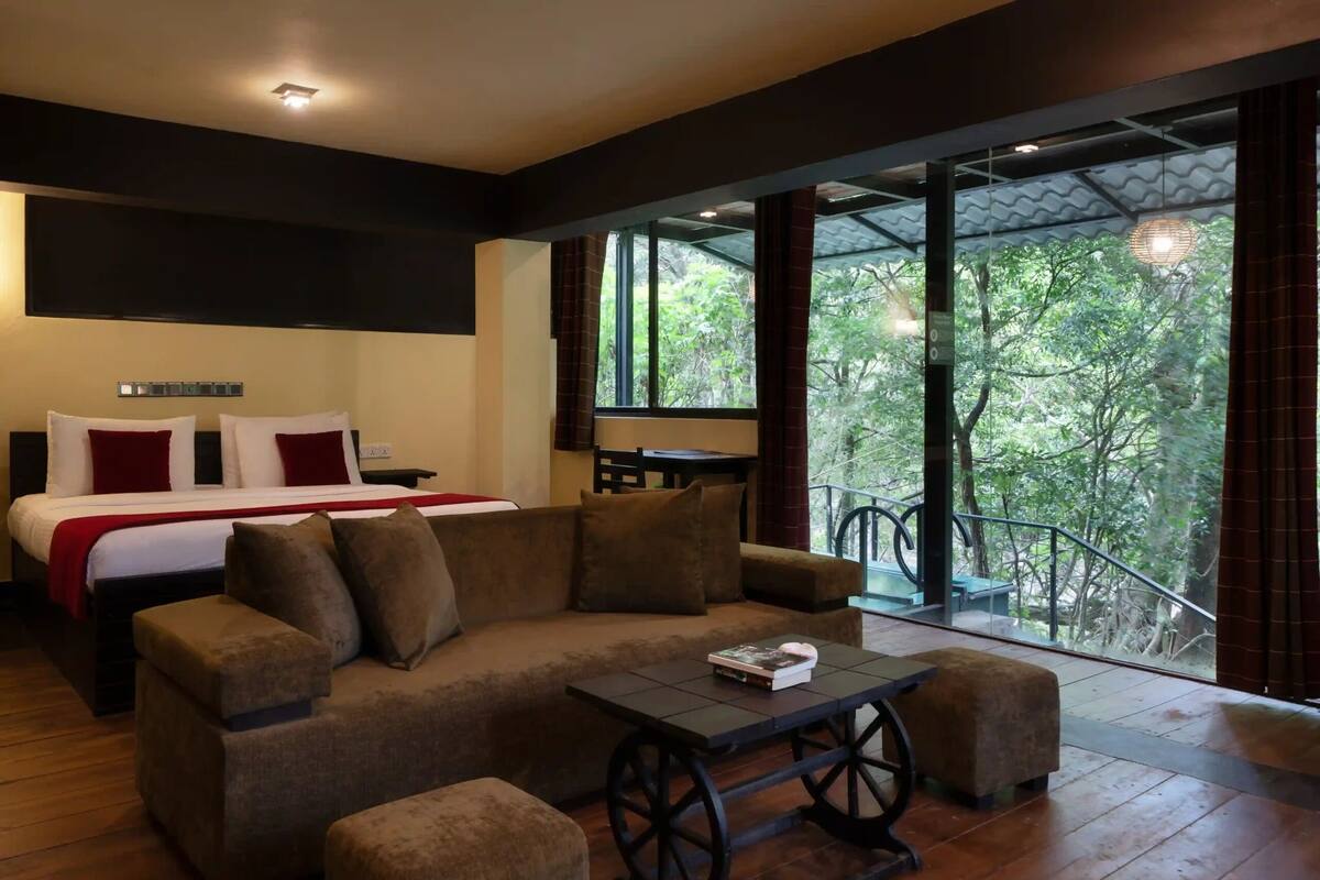 Quarto de hotel moderno com uma cama grande, um sofá, uma mesa pequena e uma varanda com vista para árvores.